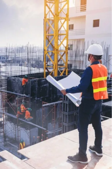 a construction worker stands in a roof, holding a blueprint and looking over a partially build building