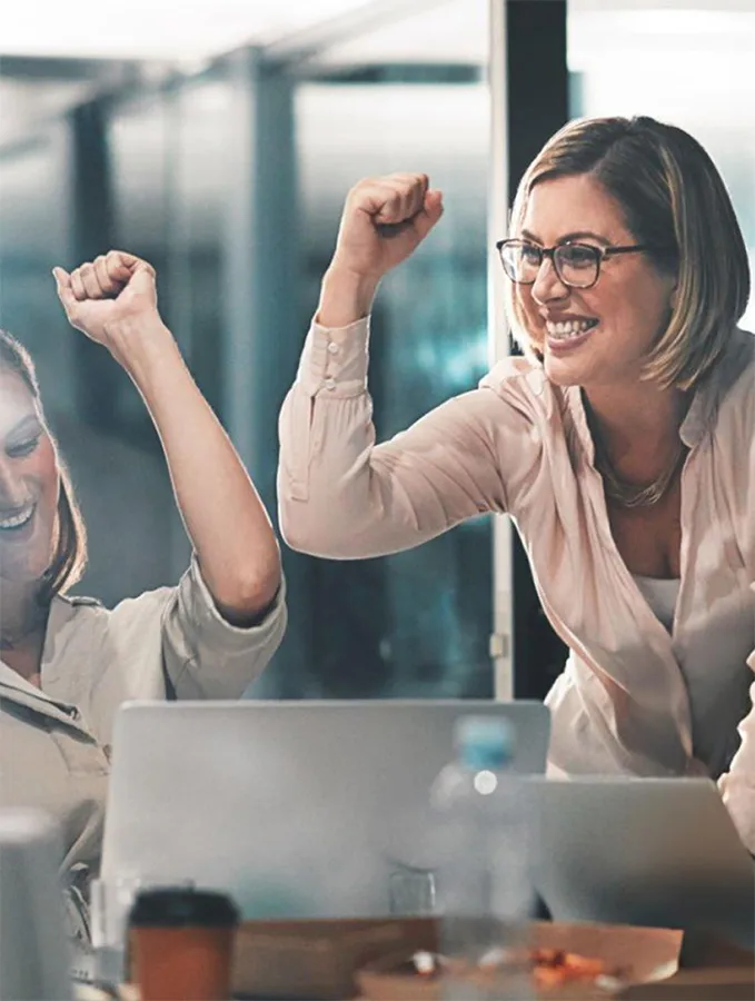 two women in an office smiling and raising their fists in celebration behind a laptop
