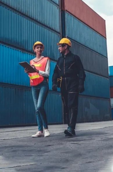two construction workers - a man and a woman - walk past shipping containers while talking to each other