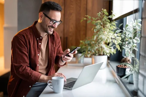 a young man sits at a clean, modern desk with his computer in front of him while smiling and looking at his phone