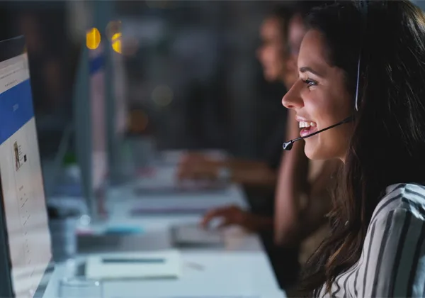 a woman sits in front of a computer with a phone headset on, smiling and talking to someone who is on the screen